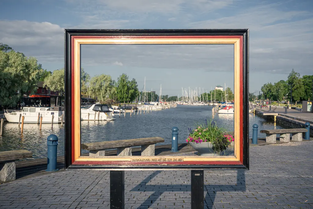 Rahmen mit Blick auf einen Hafen; Boote liegen im Wasser, umgeben von Bäumen und einer gepflasterten Promenade.