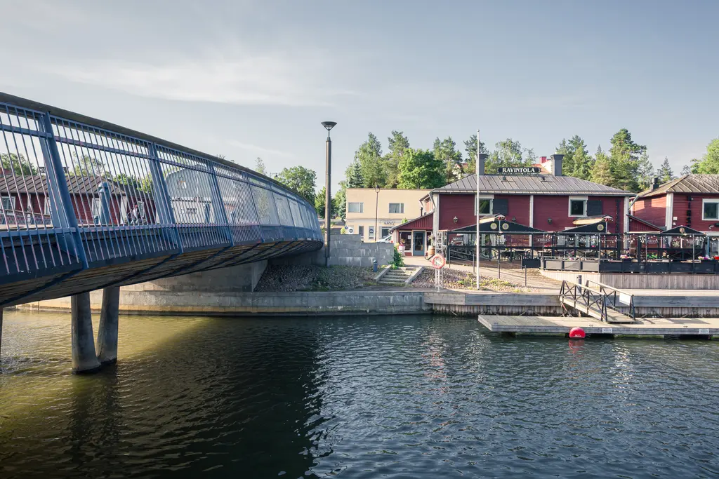 Brücke über einen Hafenkanal mit einem Restaurantgebäude und einer Terrasse im Hintergrund. Das Wasser ist ruhig und spiegelnd.