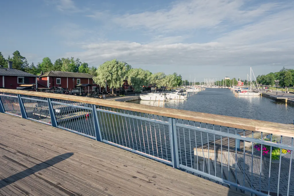 Blick auf einen Hafen mit kleinen Booten und einem Steg. Im Hintergrund sind rote Holzhäuser und grüne Bäume sichtbar.