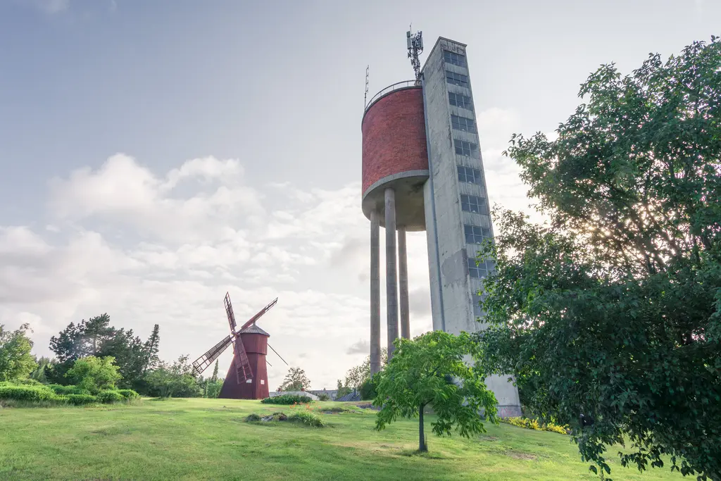 Wasserturm und Windmühle auf einer grünen Wiese, umgeben von Bäumen und leicht bewölktem Himmel.