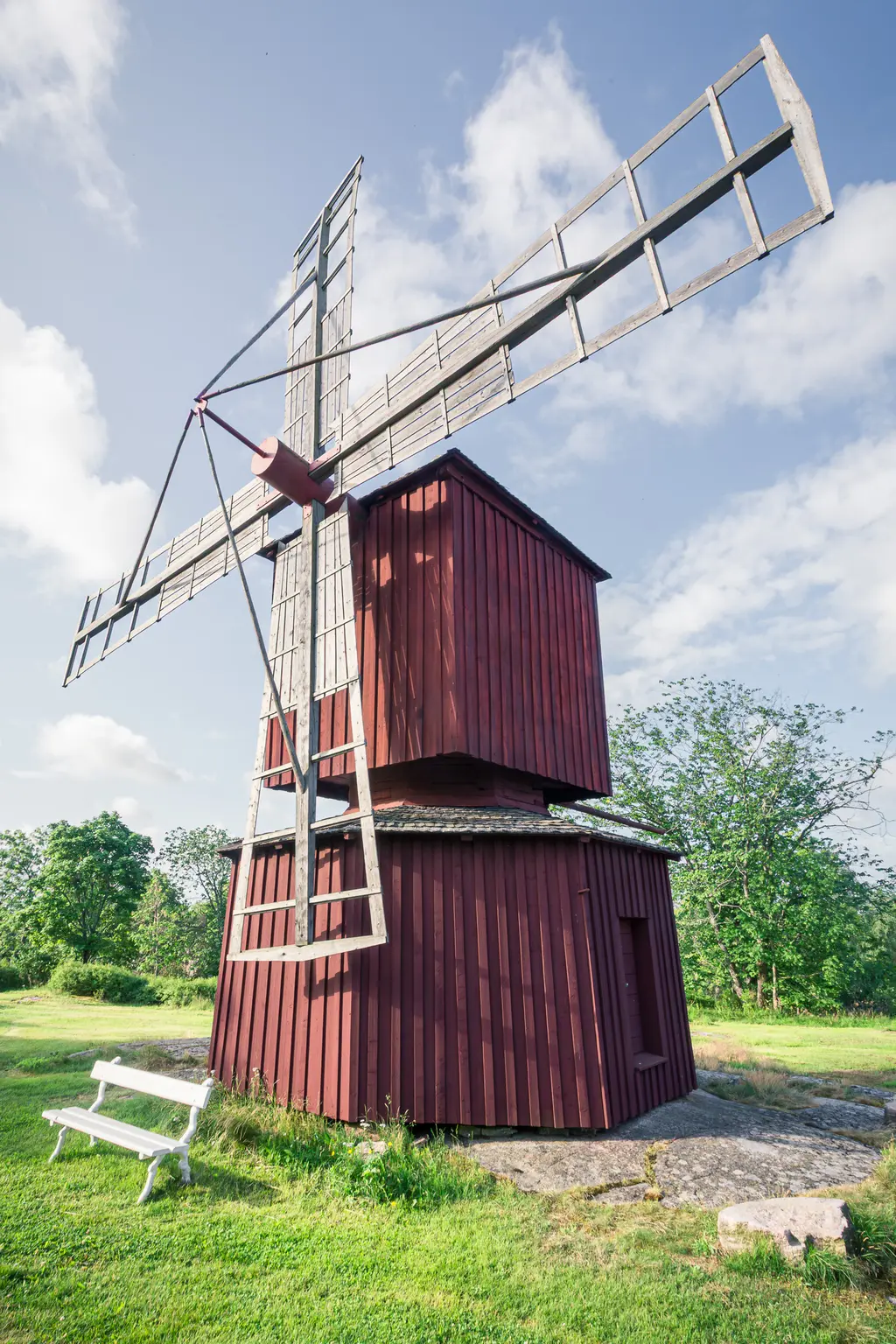 Rote Windmühle mit großen, schaufelartigen Flügeln steht auf einem grünen Rasen, umgeben von Bäumen und einem Banch.