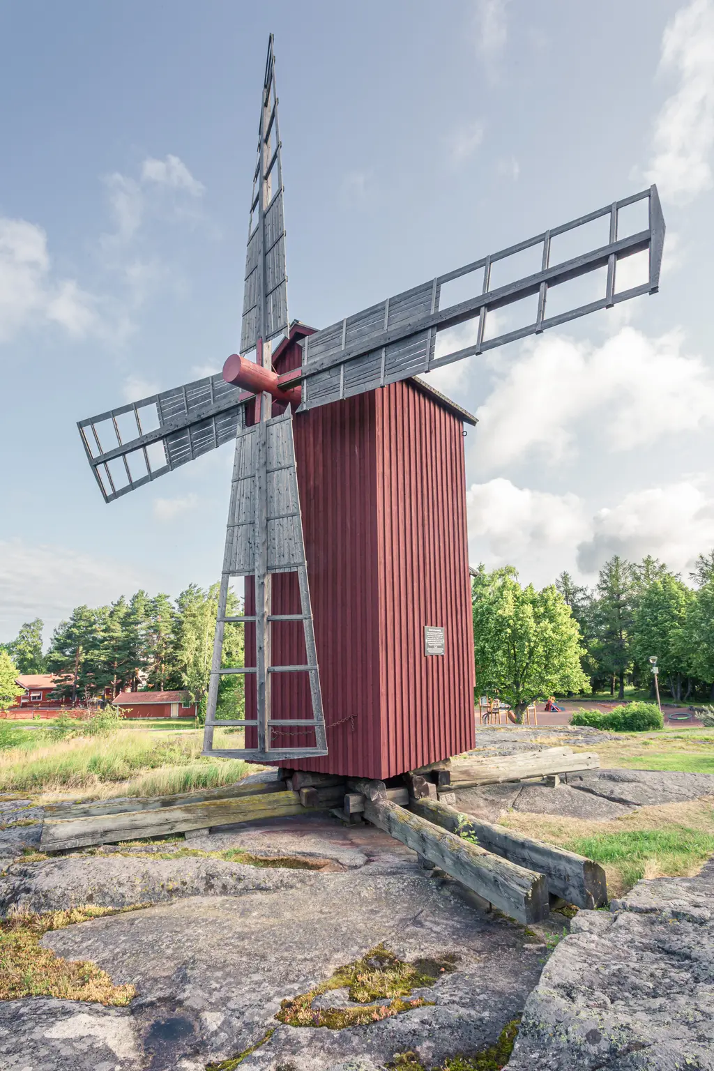 Historische Windmühle aus Holz mit großen, grauen Flügeln, umgeben von grünen Bäumen und steiniger Landschaft.