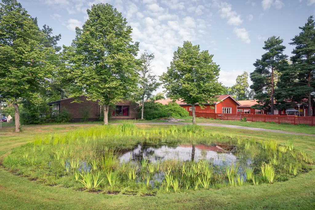 Kleiner Teich umgeben von grünen Pflanzen und Bäumen, im Hintergrund rote Holzhäuser. Der Himmel ist teils bewölkt.