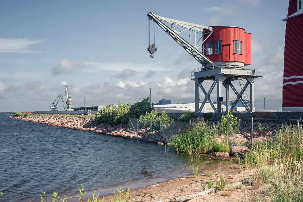 Hafenkran in Rot an einem Gewässer mit einer Uferlinie aus Steinen und Gräsern. Im Hintergrund weitere Krananlagen sichtbar.