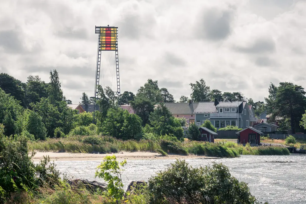 Blick auf einen Wasserbereich mit einem hohen, bunt gestreiften Turm im Hintergrund, umgeben von Bäumen und bunten Häusern.