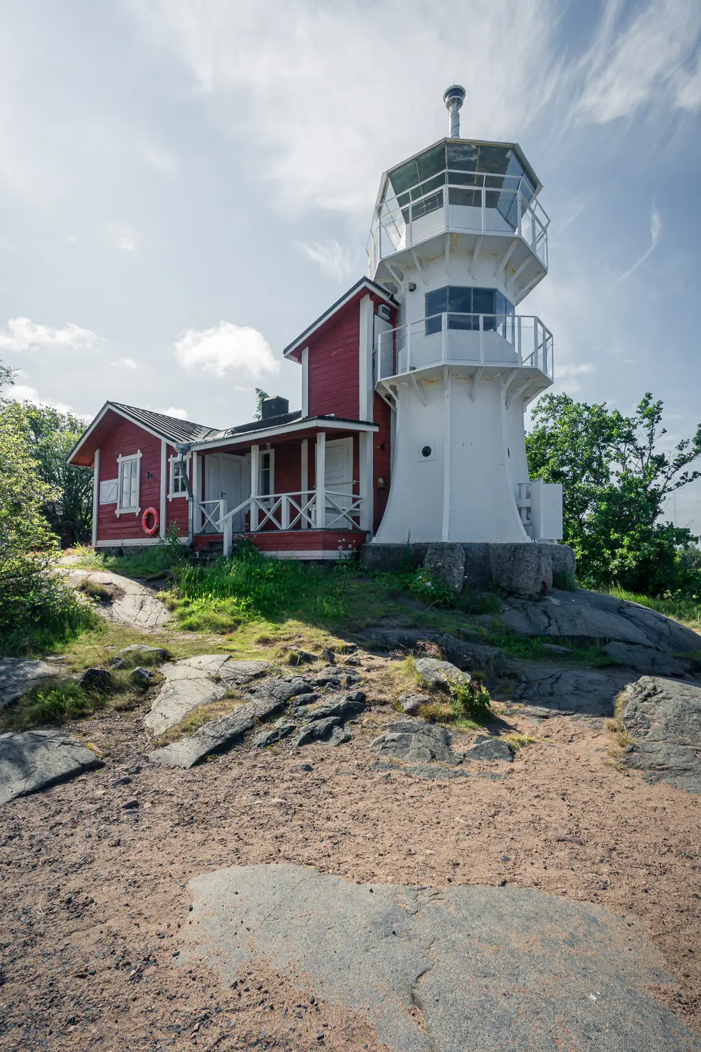 Roter Leuchtturm mit gläsernem Aussichtsturm, daneben ein kleines rotes Haus. Umgeben von felsigem Boden und grüner Vegetation.