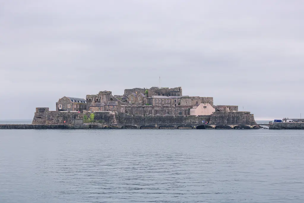 A coastal fortress with stone walls and various buildings, surrounded by calm waters and a cloudy sky.