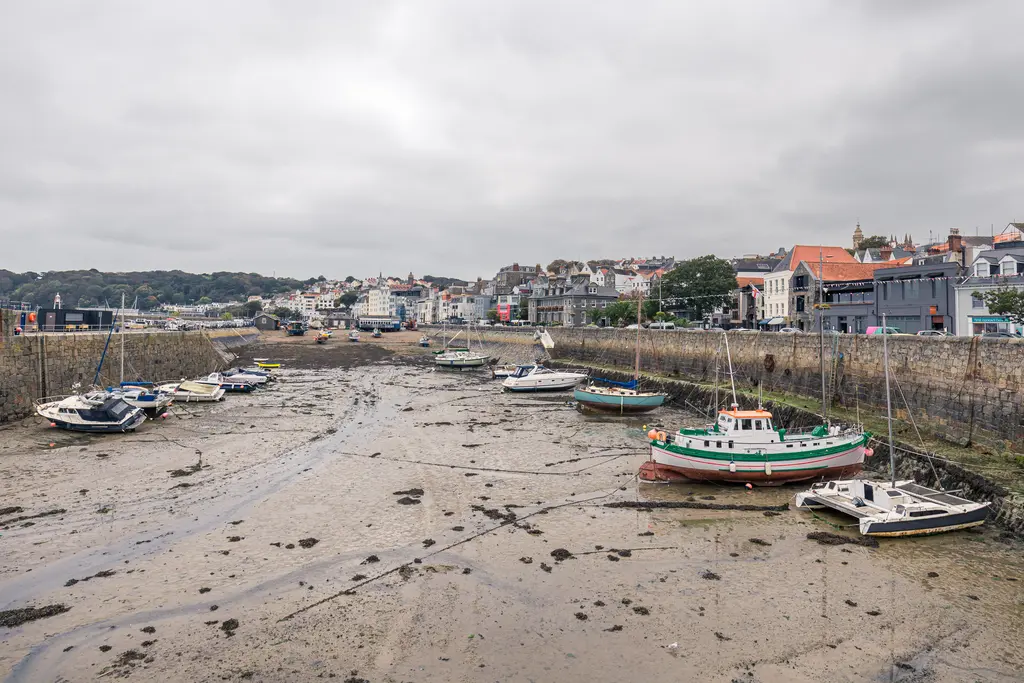 A harbor scene with boats resting on mudflats at low tide, surrounded by colorful buildings and a cloudy sky.