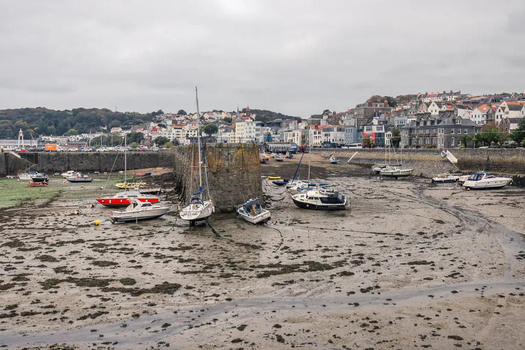 Boats resting on mudflats at low tide with coastal town buildings in the background under a cloudy sky.