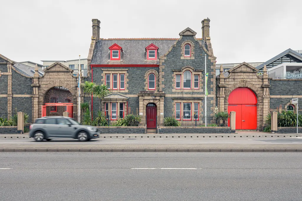 Historisches, graues Gebäude mit roten Akzenten und Wohnfenstern, daneben ein Parkplatz und eine Straße mit einem passierenden Auto.