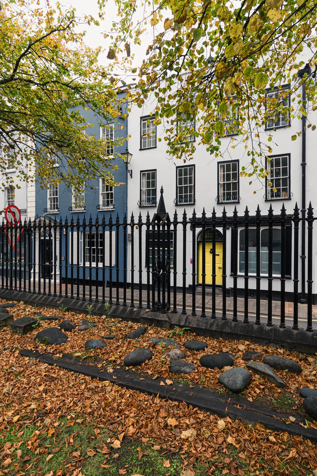Bunte Häuserfassaden in Blau und Weiß, mit einem gelben Eingang. Vor einem eisernen Zaun liegen orange-braune Herbstblätter und Steine.