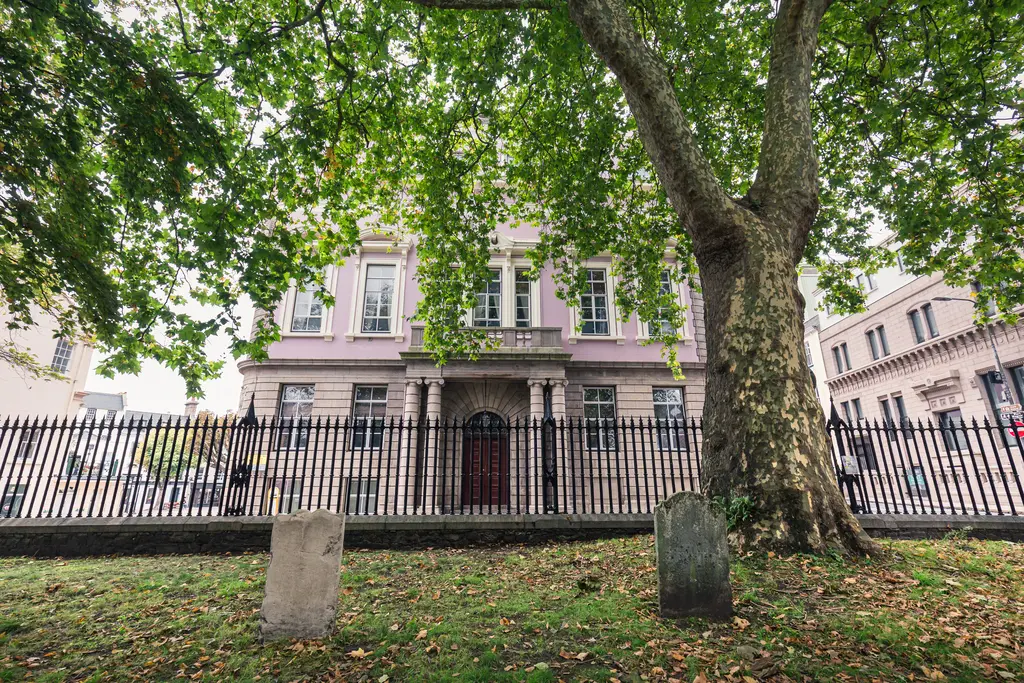 Historisches Gebäude mit rosa Fassade, umgeben von einem Zaun. Im Vordergrund steht ein großer Baum und einige Grabsteine auf grünem Rasen.