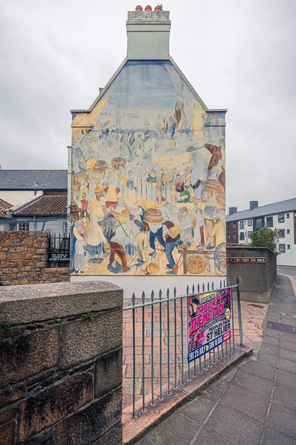 Wandgemälde an einem Gebäude, das eine lebhafte Marktszene darstellt. Im Vordergrund ist ein Zaun mit einem Plakat für einen Themenpark sichtbar.
