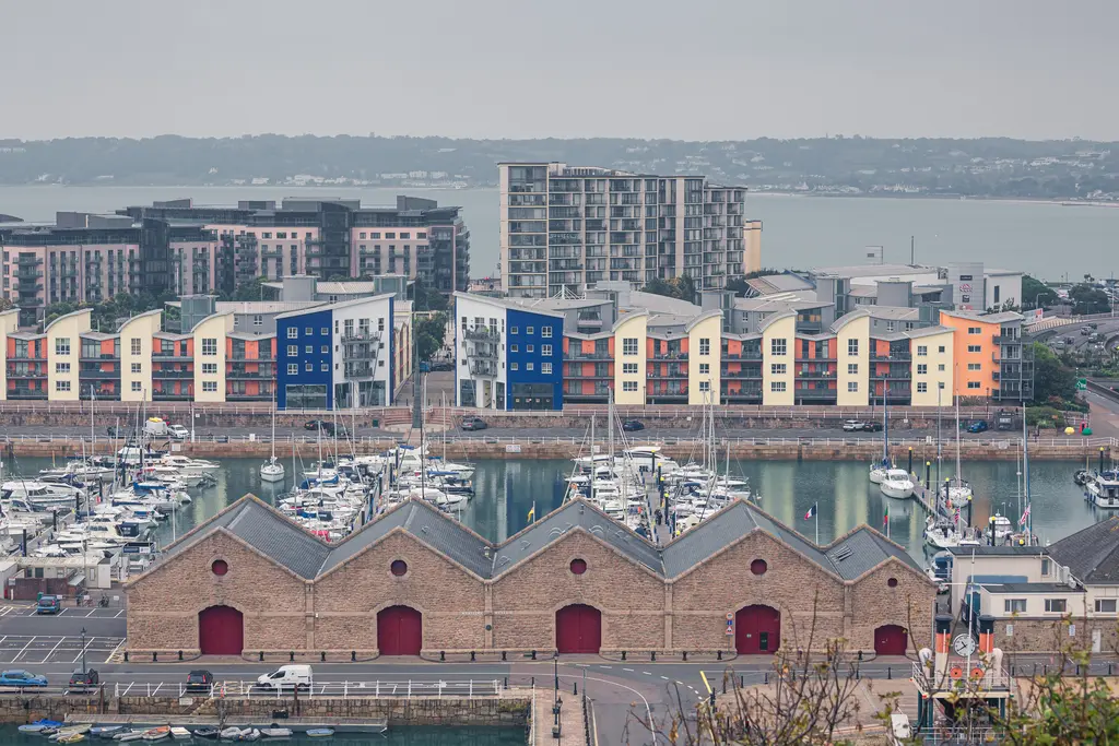 Moderne Wohnhäuser mit bunten Fassaden neben einem Hafen mit Yachten und historischen Lagerhäusern im Vordergrund.