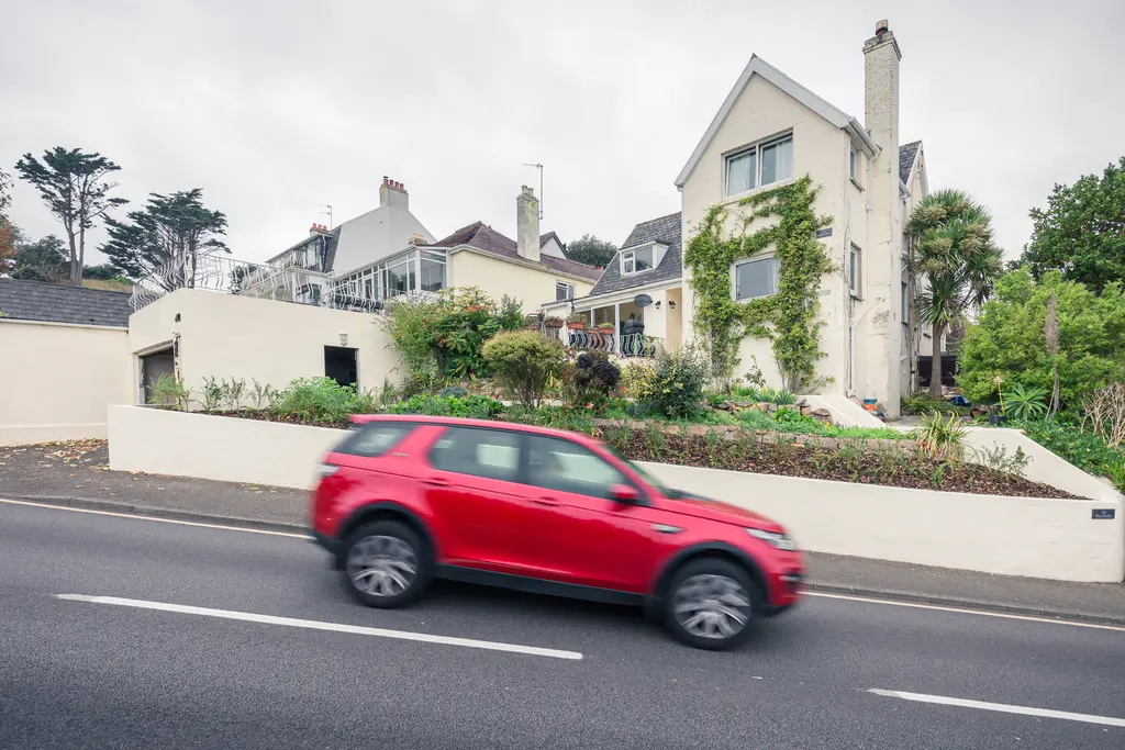 Rotes Auto fährt an einem zweigeschossigen, weißen Haus mit grüner Bepflanzung vorbei. Im Hintergrund sind weitere Gebäude und Bäume sichtbar.