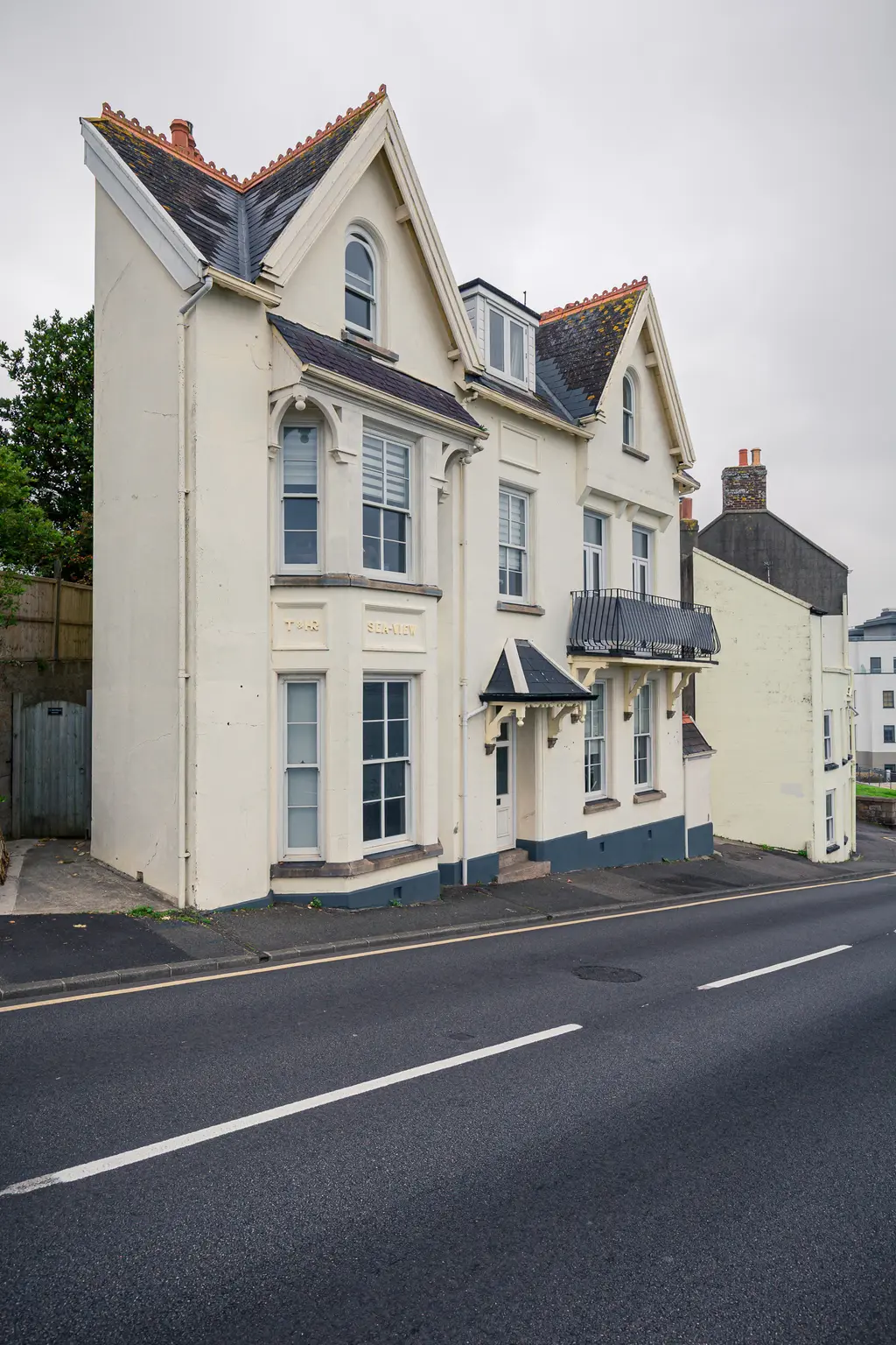 Dreistöckiges, helles Wohngebäude mit Satteldach und Fenstern im oberen Bereich. Eine kleine Veranda und eine gepflasterte Auffahrt sind sichtbar.