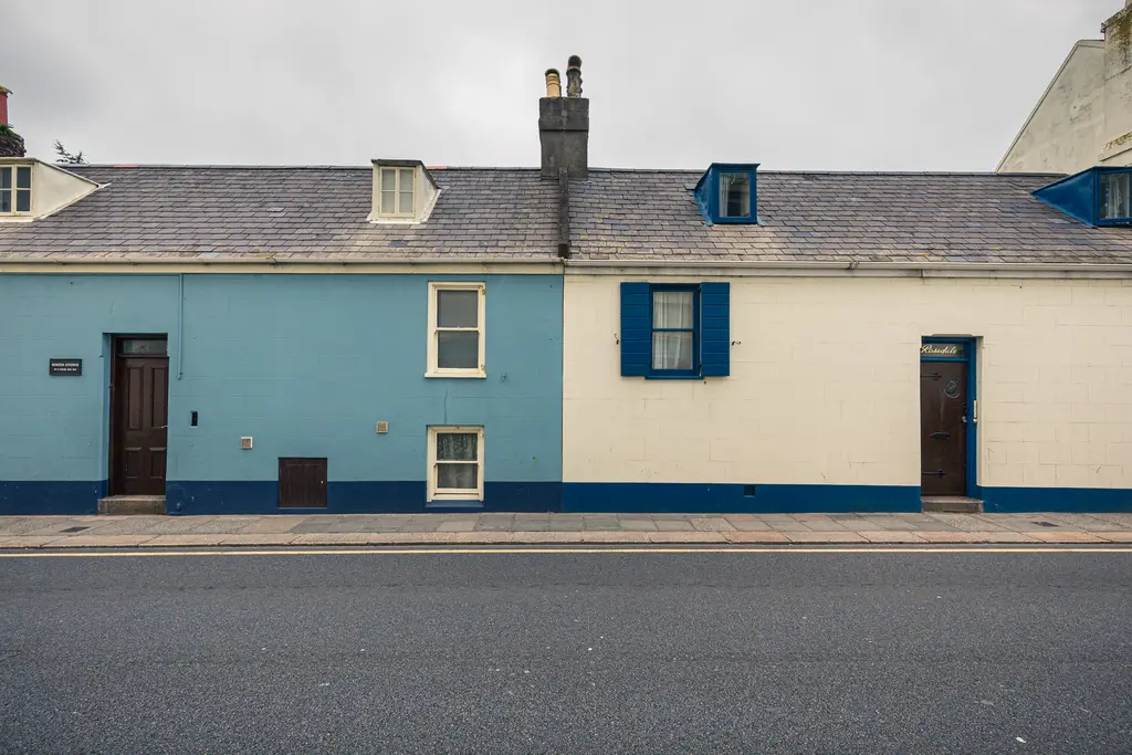 Zwei nebeneinanderstehende Häuser mit unterschiedlichen Farben: links in Blau, rechts in hellem Weiß mit blauen Fensterläden.
