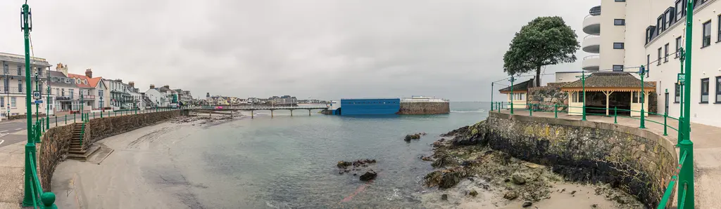 Küstenansicht mit flachem Wasser, felsigem Ufer und Sandstrand. Links sind Gebäude und Laternen sichtbar. Ein blauer Container liegt im Wasser.