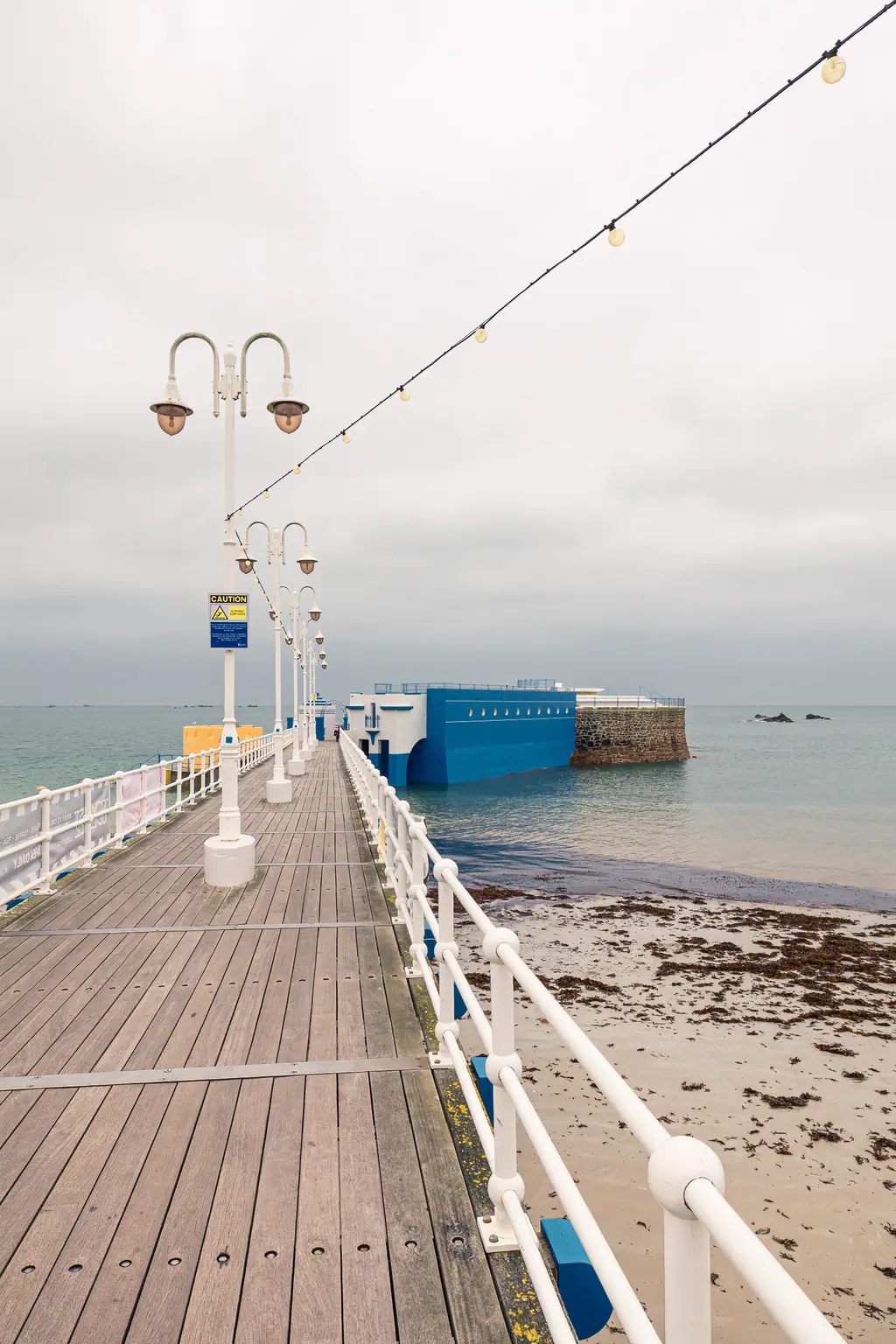 Holzsteg mit Beleuchtung, der in Richtung eines blauen Gebäudes am Wasser führt. Der Himmel ist bewölkt, der Strand ist sichtbar.
