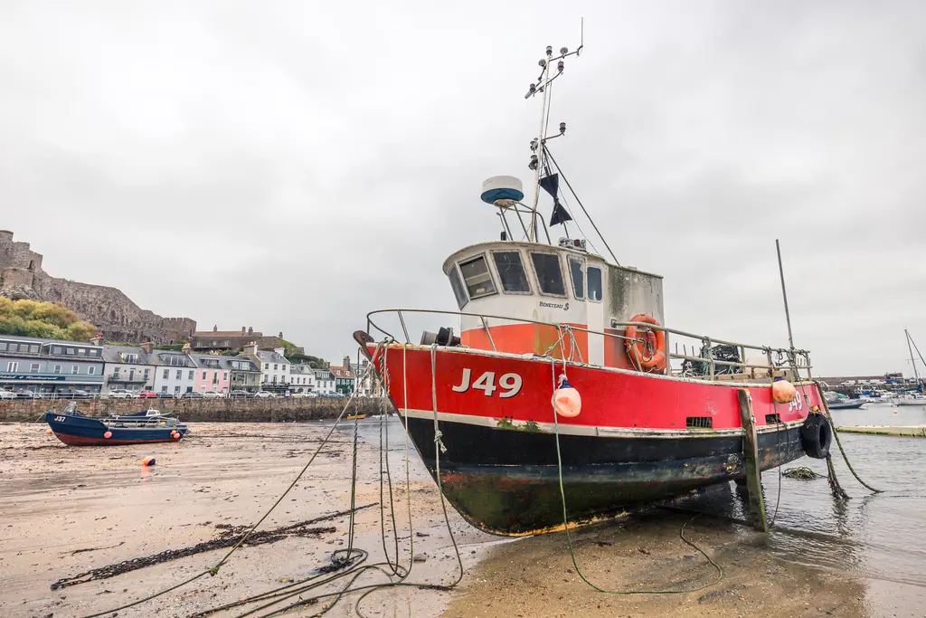 Fischerboot mit rotem Rumpf und der Kennung J49, auf trockenem Sand angelandet. Im Hintergrund sind Häuser und ein Hügel zu sehen.