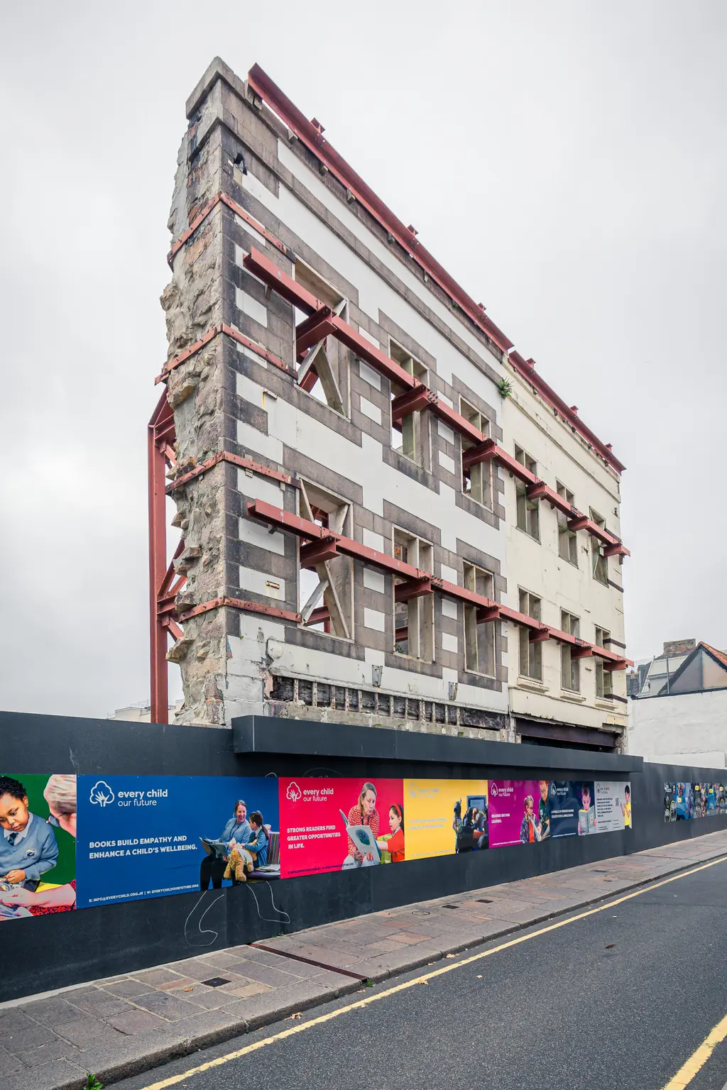 Rohbau mit sichtbaren Stahlträgern und einer teilweise abgerissenen Wand. Vor dem Gebäude verläuft ein Bauzaun mit bunten Werbetafeln.