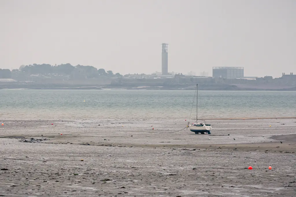 Boot auf dem trockenen Sandstrand, umgeben von flachem Wasser und bunten Bojen im Hintergrund. Eine Industrieanlage ist sichtbar.