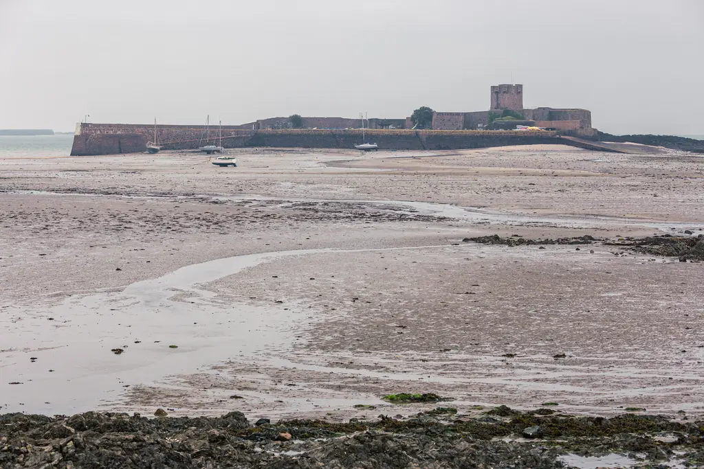 Burg auf einer Halbinsel, umgeben von trockenem, schlammigem Boden. Kleine Boote liegen im Flachwasser. Der Himmel ist bewölkt.