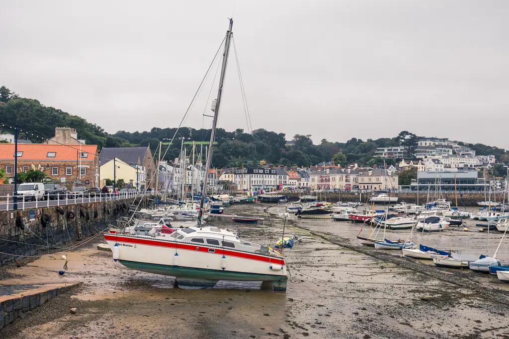 Hafen mit mehreren Booten im Trockenen, darunter ein größeres Segelboot. Im Hintergrund sind bunte Häuser und bewaldete Hügel sichtbar.