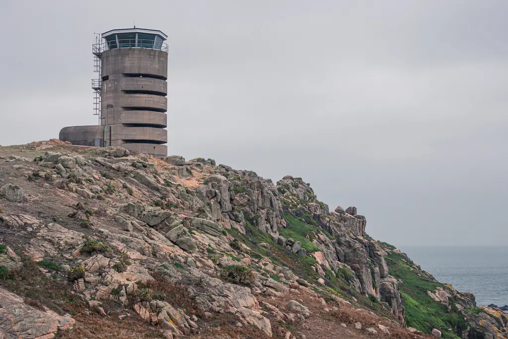 Beobachtungsturm auf einer felsigen Küstenlandschaft, umgeben von grünem Gras und steinigen Ausläufern, unter bewölktem Himmel.