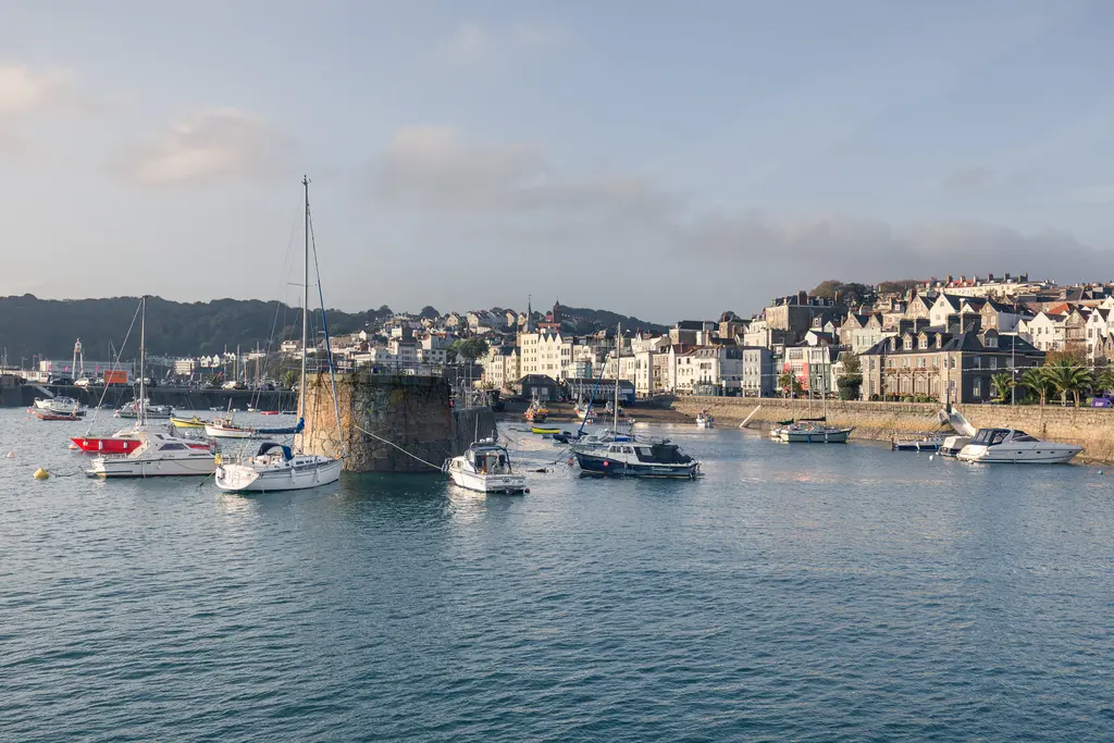 A picturesque harbor with sailing boats anchored near a stone pier, surrounded by charming coastal buildings and green hills.