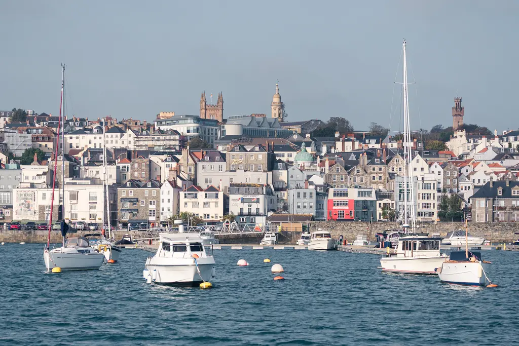 A coastal scene featuring various boats anchored in calm water, with colorful buildings lining the shore and hills in the background.