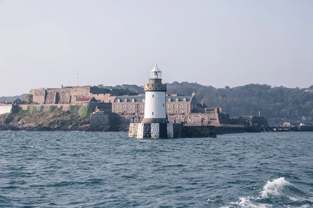 A lighthouse stands on a rocky foundation with a historic fort in the background, surrounded by calm waters.