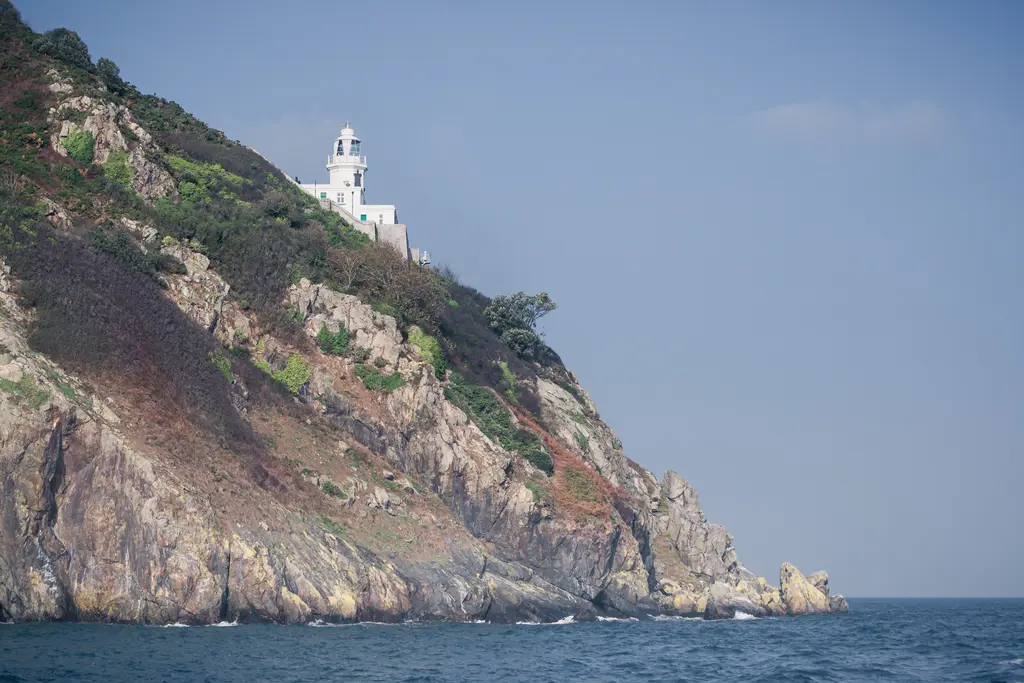 A lighthouse stands atop a rocky cliff, surrounded by lush greenery, overlooking calm blue waters under a clear sky.