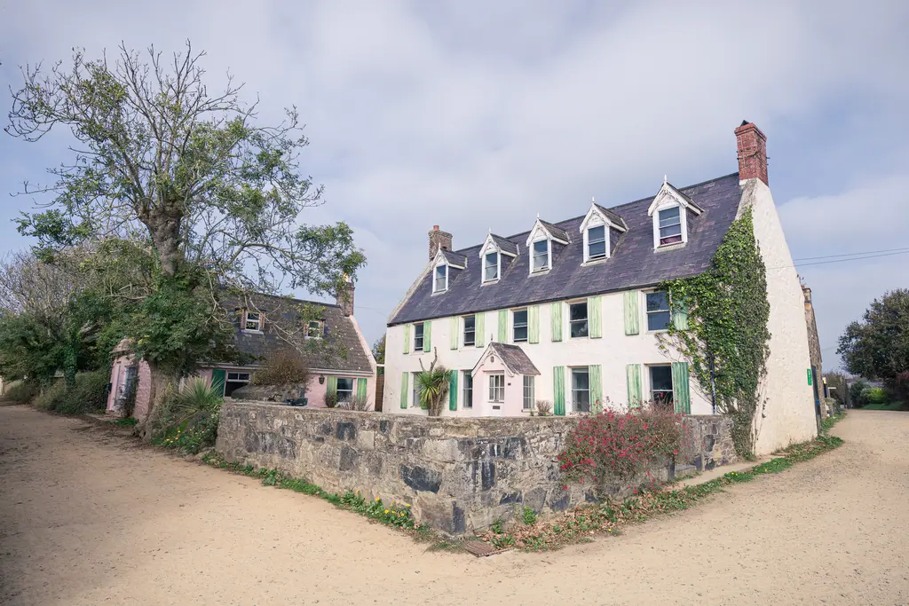 A traditional stone house with a slate roof and green shutters, surrounded by trees and a sandy path.