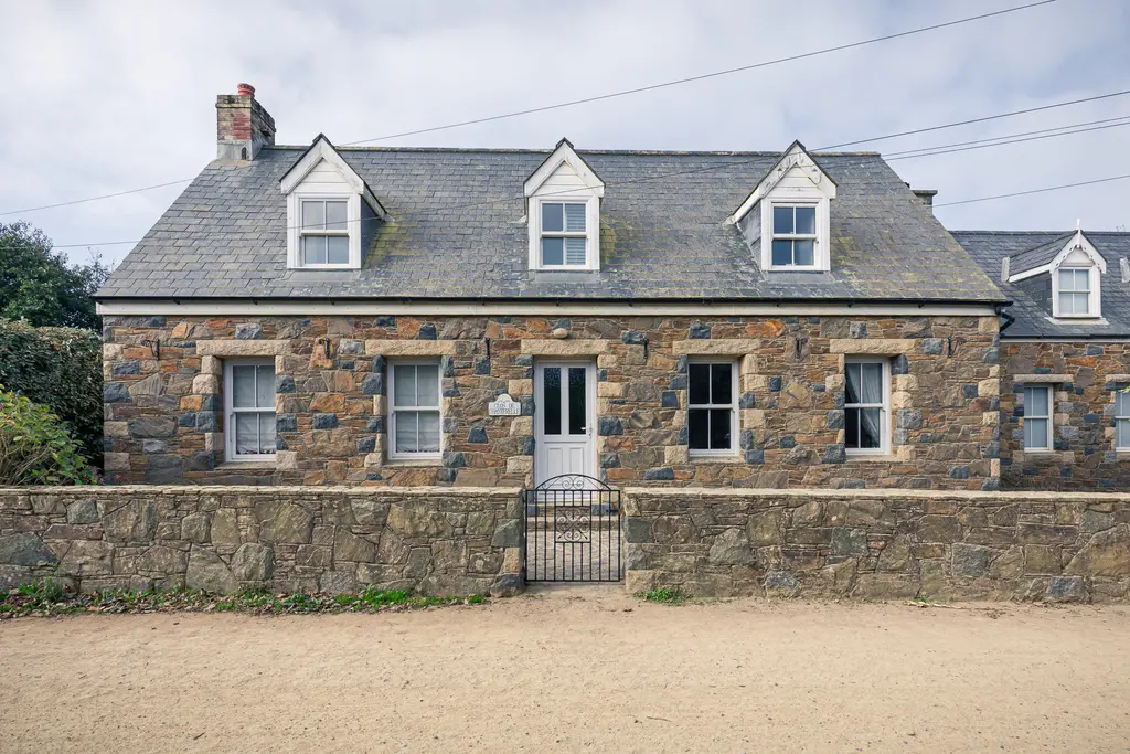 A stone cottage with a slate roof and white door, surrounded by a low stone wall and garden. A quaint architectural feature on Sark.