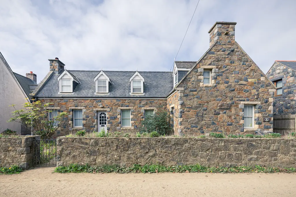 A charming stone house with multiple gabled windows, surrounded by greenery and a low stone wall, under a cloudy sky.