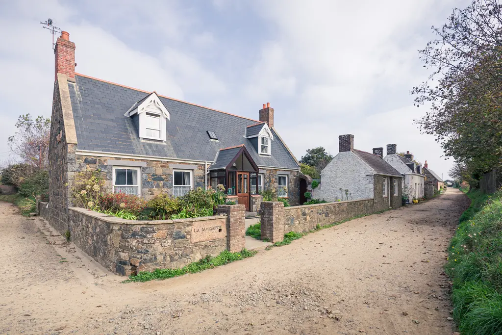 A charming stone cottage with a slate roof on a sandy road, surrounded by greenery and additional quaint houses.