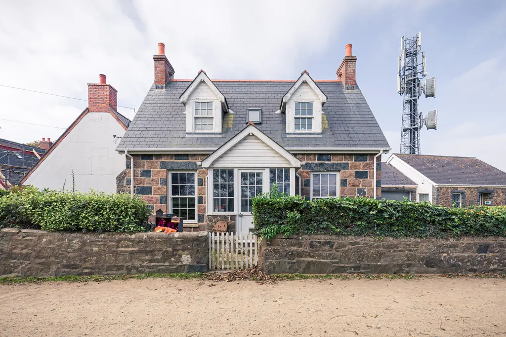 A quaint stone cottage with a pitched roof, surrounded by greenery and a low stone wall, with a telecommunications tower in the background.