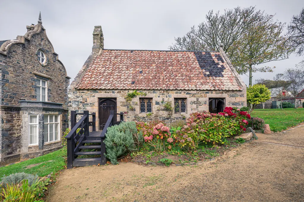 A charming stone cottage with a tiled roof, surrounded by colorful flowers and greenery, near a path in a rural setting.