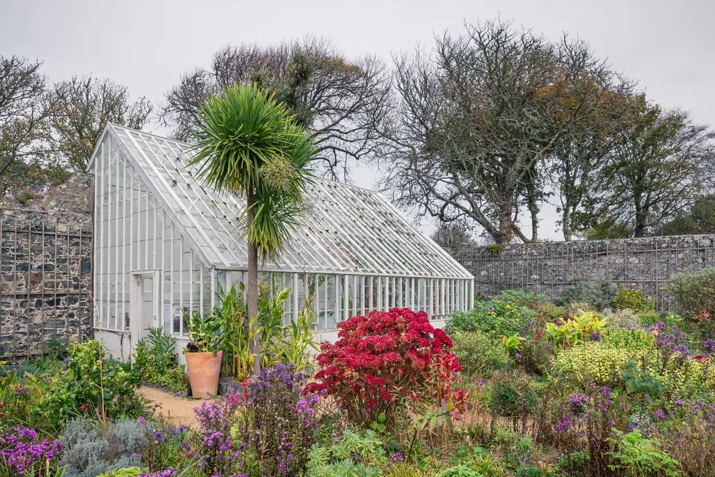A glass greenhouse surrounded by colorful flowers and lush greenery, with leafless trees in the background on a cloudy day.