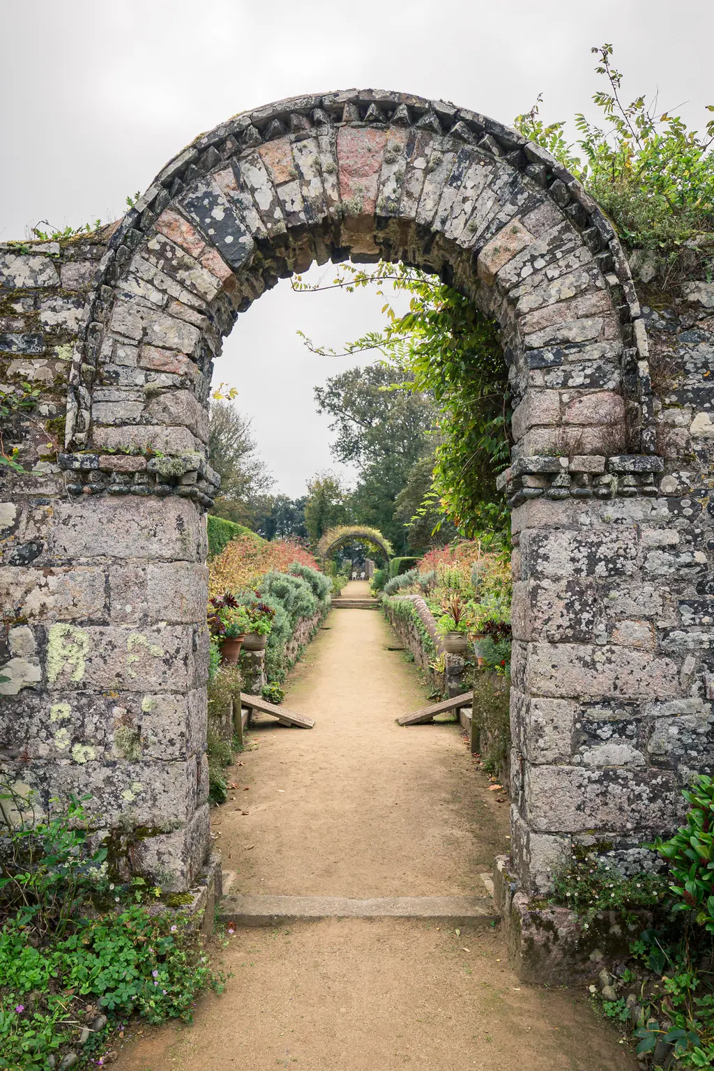 A stone archway frames a garden path lined with potted plants, leading toward lush greenery in a serene setting.