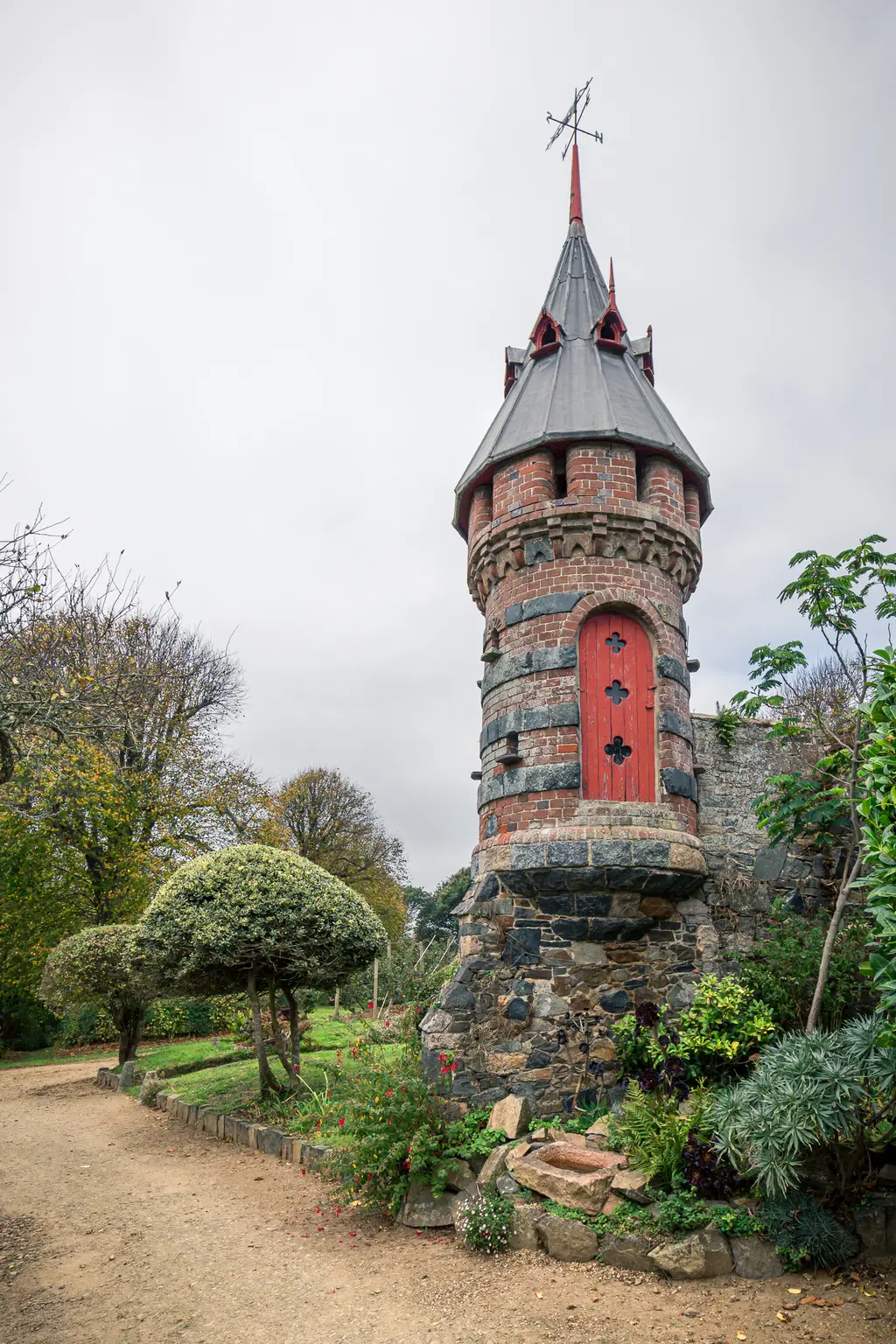 A decorative stone tower with a red door and spire, surrounded by lush greenery and a walking path.