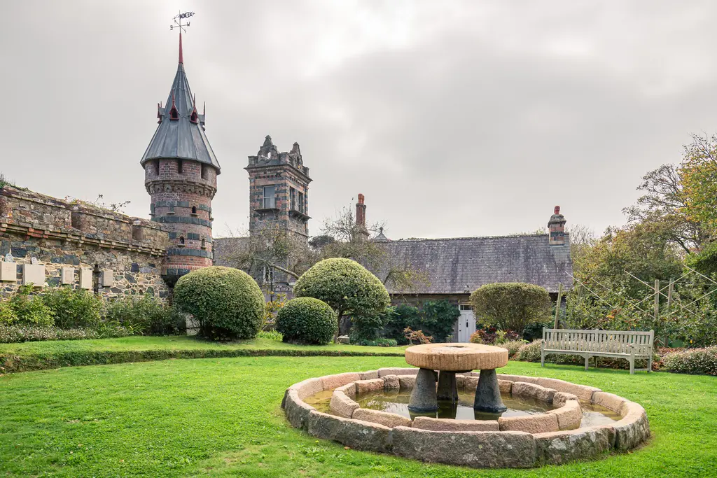 A historic castle garden with a stone fountain, manicured lawns, and a tower with a weathervane under a cloudy sky.