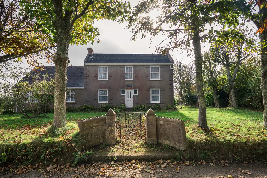 A charming two-story house surrounded by trees and a grassy lawn, featuring a gated entrance on a quiet lane.