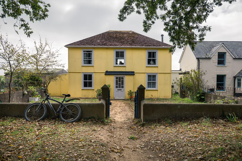 A yellow house with a front garden, surrounded by trees, and a bike parked near the entrance path.