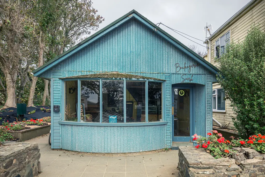 A charming blue cottage with large windows, surrounded by vibrant flowers and greenery, showcasing island architecture.