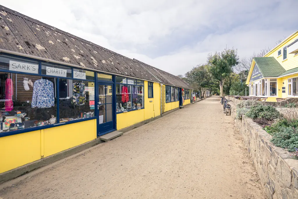 A sandy path lined with colorful shops, including a charity shop, surrounded by greenery and quaint buildings.