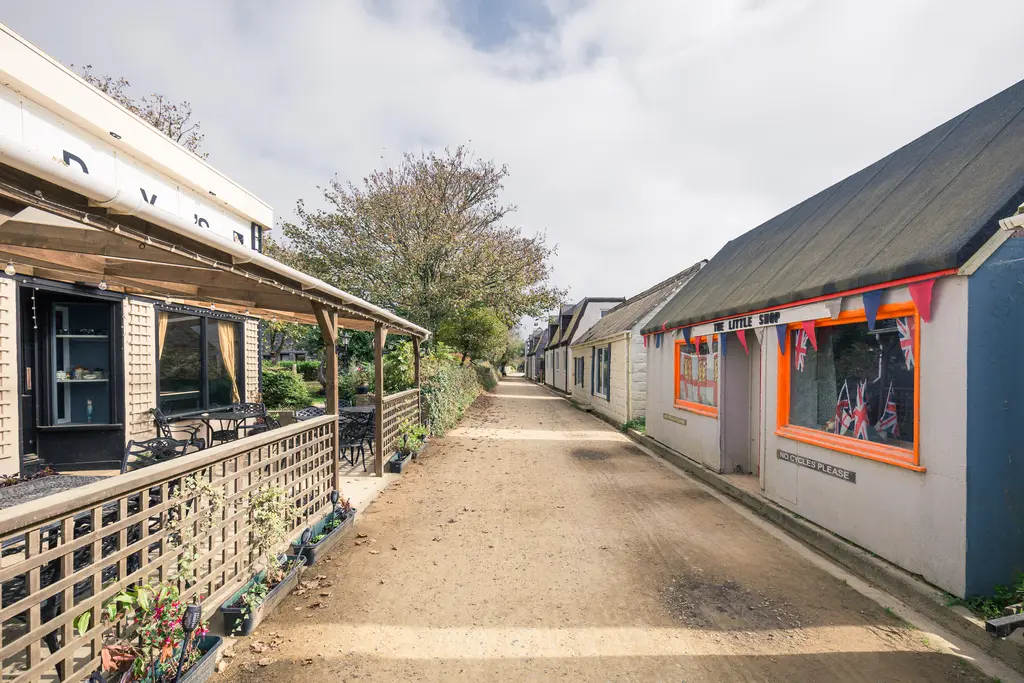 A narrow village path lined with quaint shops and greenery, leading through a charming coastal community on a cloudy day.