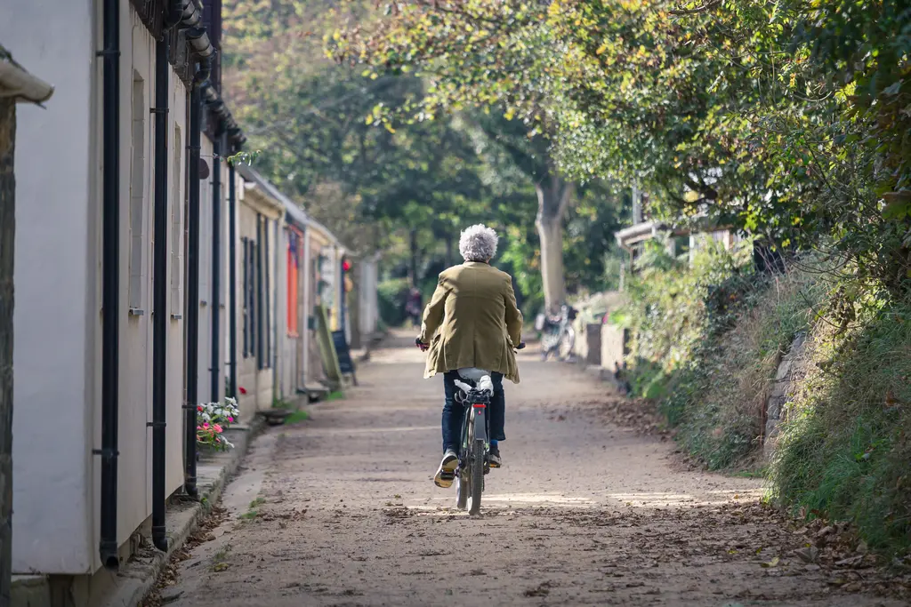 A person cycling down a tree-lined path next to quaint cottages, surrounded by greenery and fall foliage.