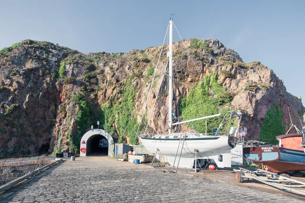 A peaceful harbor scene with a sailboat on a cobblestone path, surrounded by rocky cliffs and greenery, and a dark tunnel entrance.